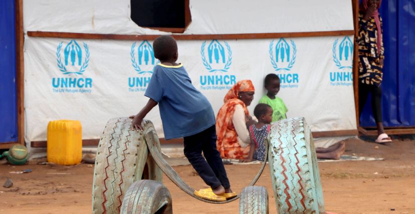 أطفال لاجئون سودانيون وأمهم أمام خيمة بها شعار UNHCR
